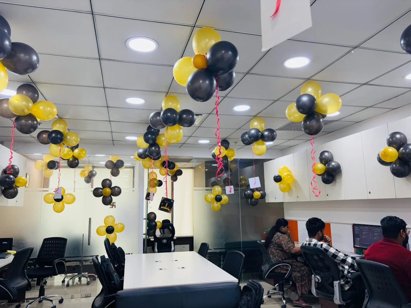 Clusters of black and yellow balloons hung across the office ceiling with Celebrating the 1st Anniversary cards dangling from them, three colleagues working at their desks below.