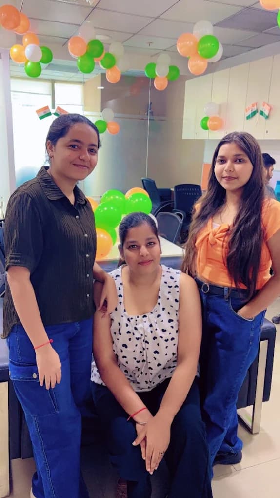Three colleagues, one seated on the sofa arm, in front of clusters of orange, white and green balloons in the office for Independence Day 2024.