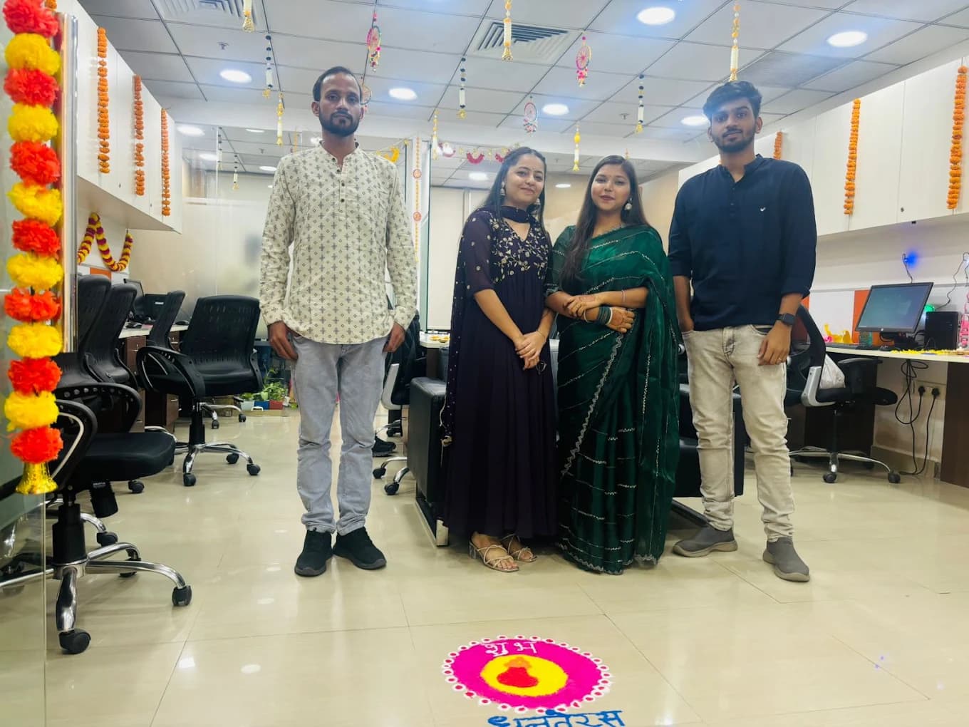 Four colleagues in a kurta, a purple dress, a green saree and a navy shirt standing between the desks under marigold strings, the Shubh Dhanteras rangoli on the floor in front of them.
