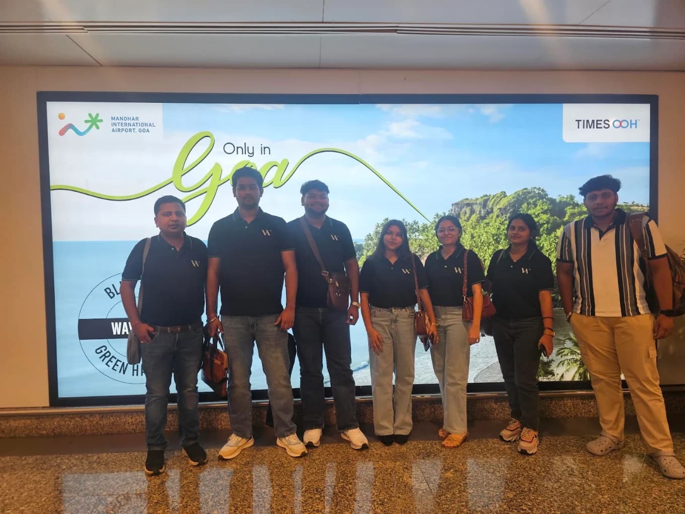 Seven team members, six in black Web Spider polos, lined up in front of the Only in Goa hoarding at Manohar International Airport after landing.