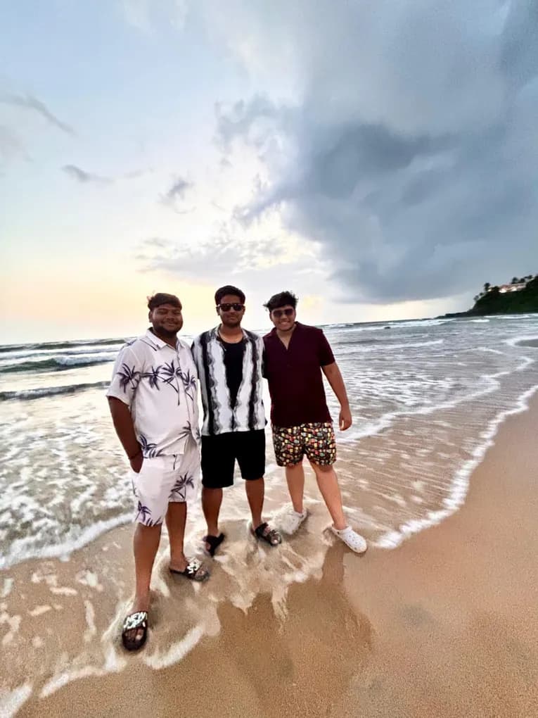 Three colleagues in beach shirts standing in the surf on a Goa beach at dusk, storm clouds over the sea.