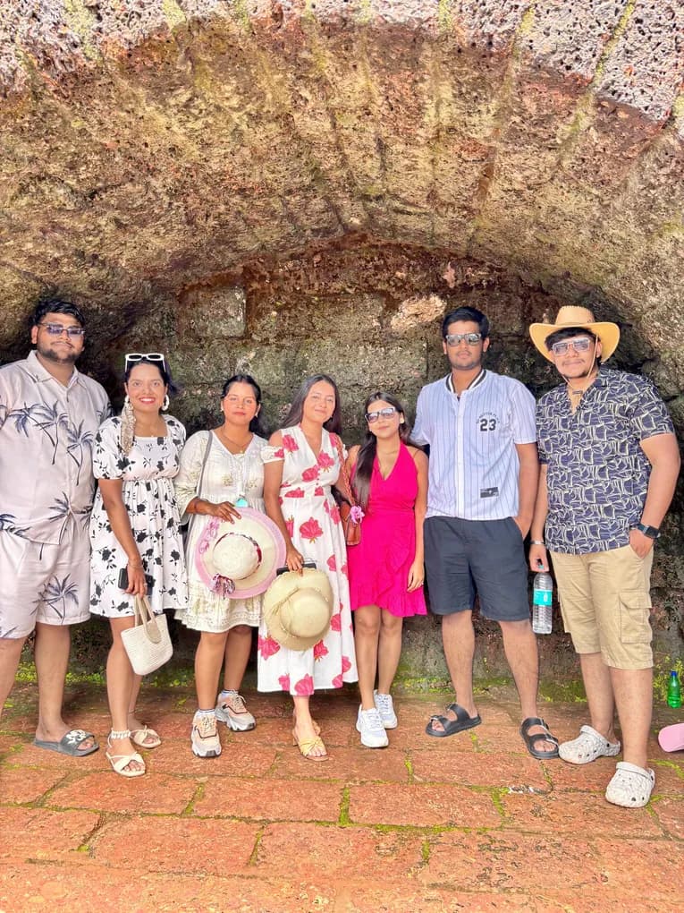 Seven team members in beach shirts and sundresses, two holding straw hats, under a laterite stone arch at the fort in Goa.