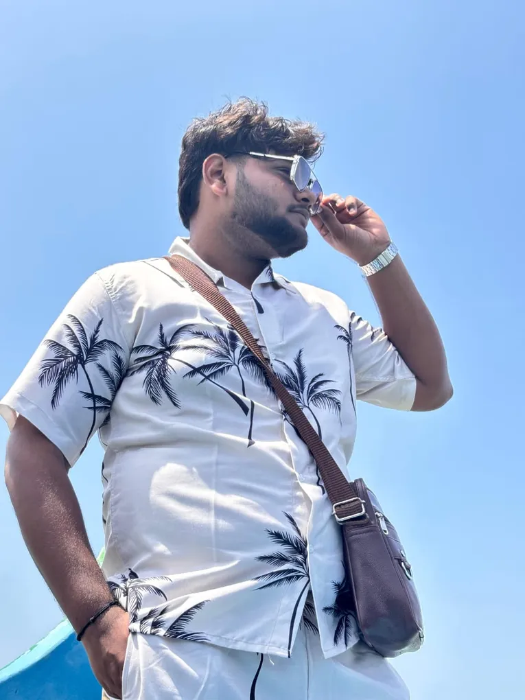 A colleague in a palm-print shirt and sunglasses against a clear blue sky, a leather sling bag over one shoulder.