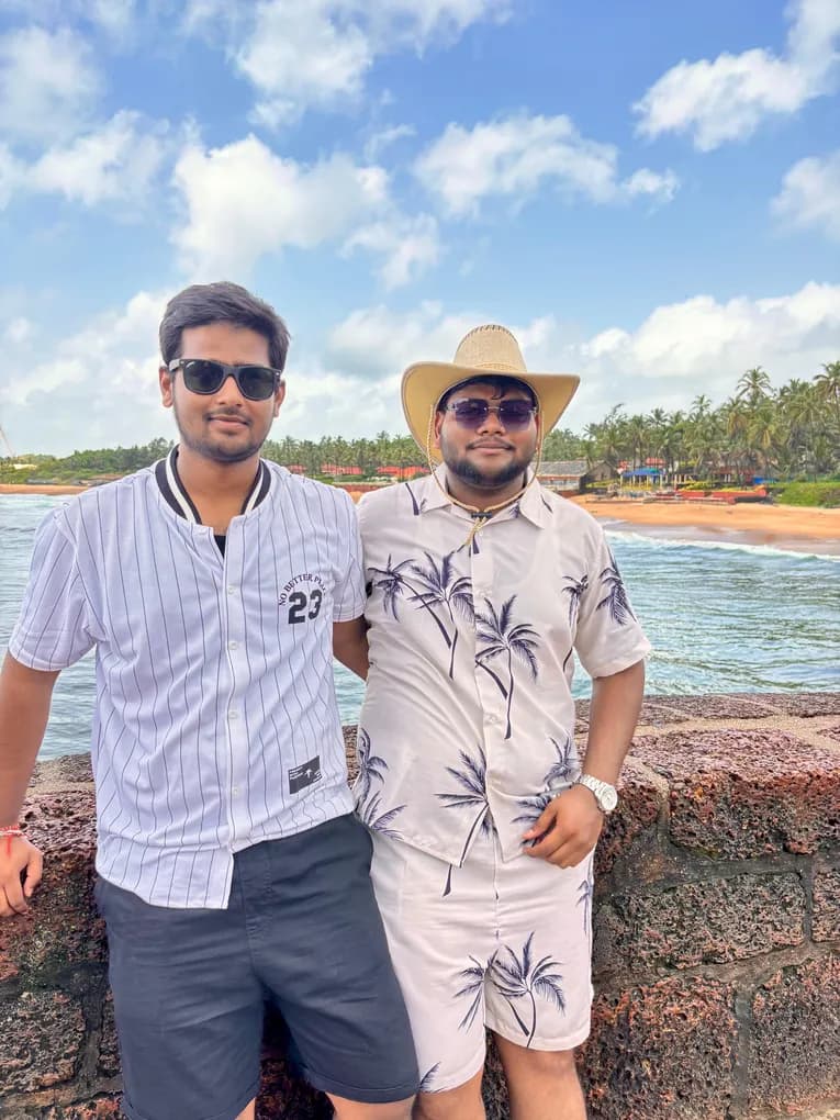 Two colleagues, one in a pinstriped jersey and one in a straw hat and palm-print shirt, leaning on the fort wall above the beach in Goa.