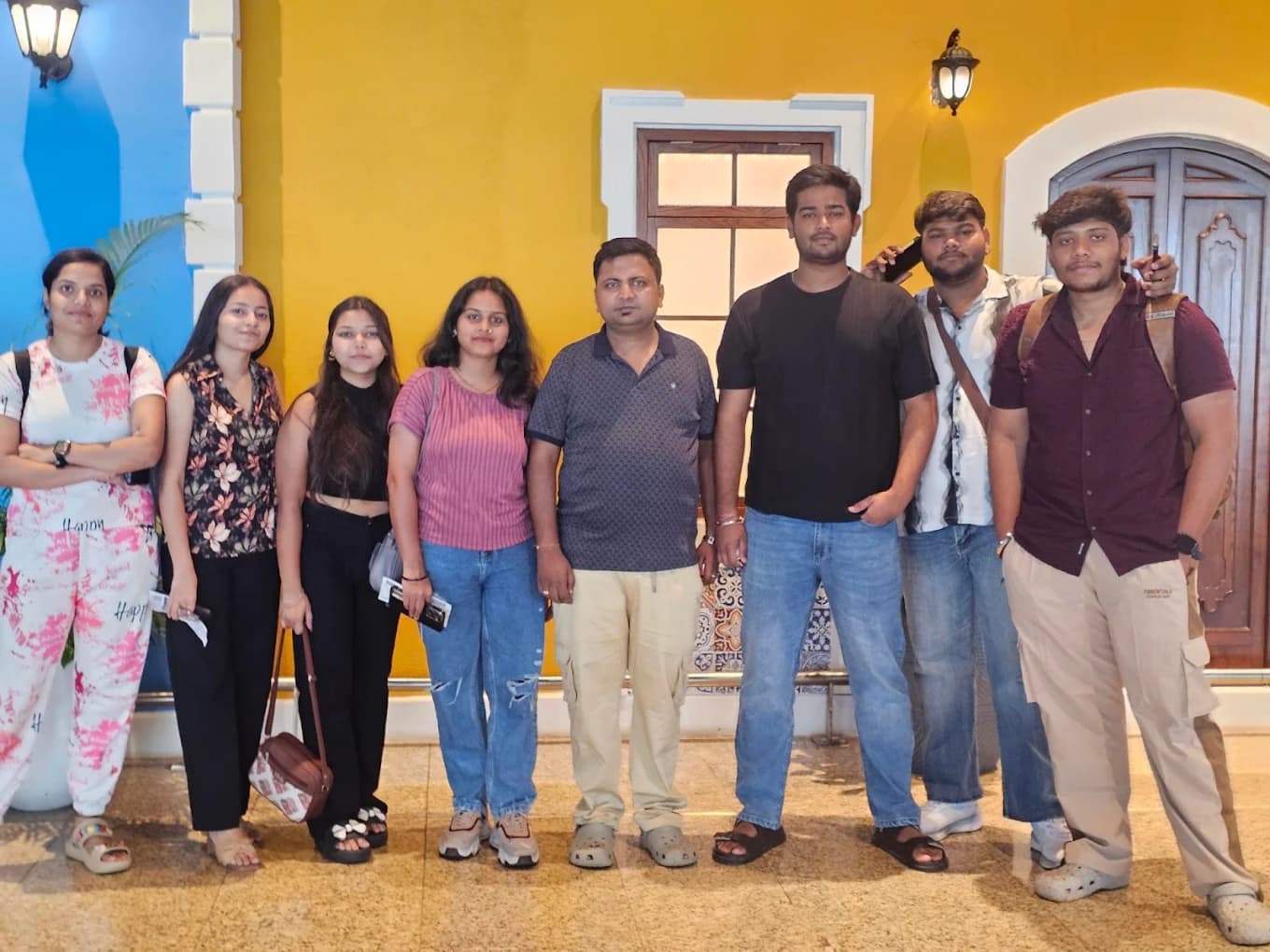 Eight team members lined up against a yellow Portuguese-style wall with blue trim, lantern sconces and a wooden door in Goa.