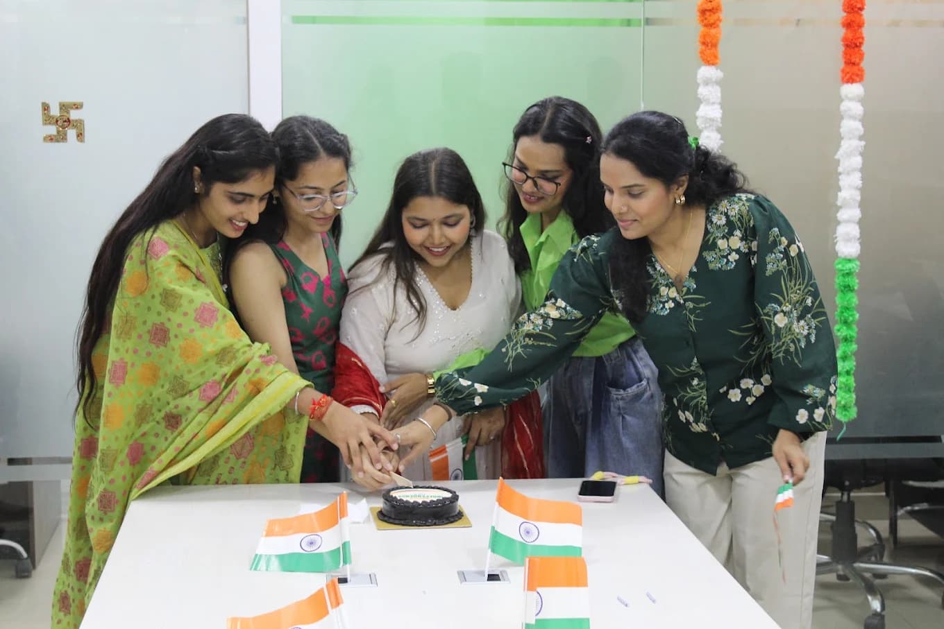 Five colleagues in green, white and red cutting a cake together on the boardroom table, small Indian flags standing around it.