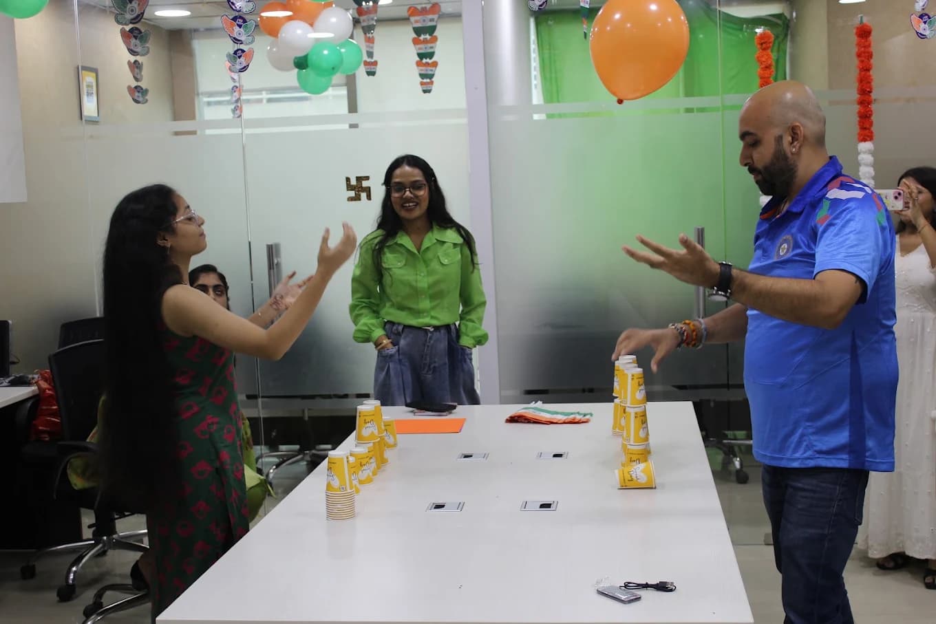 Two colleagues racing to stack paper cups at either end of the boardroom table, one in a blue India cricket jersey, a colleague in a green shirt between them keeping score.