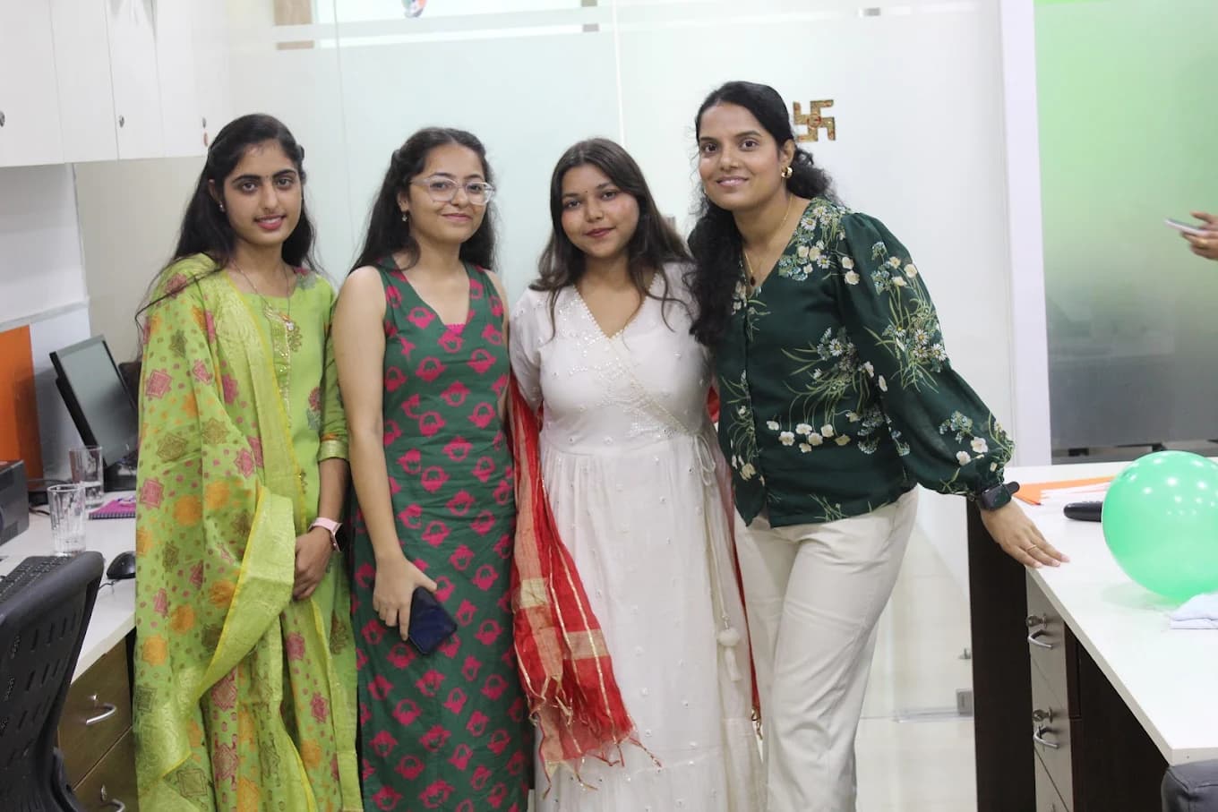 Four colleagues in green, pink and white ethnic wear standing between the desks, a green balloon beside them, for Independence Day 2025.
