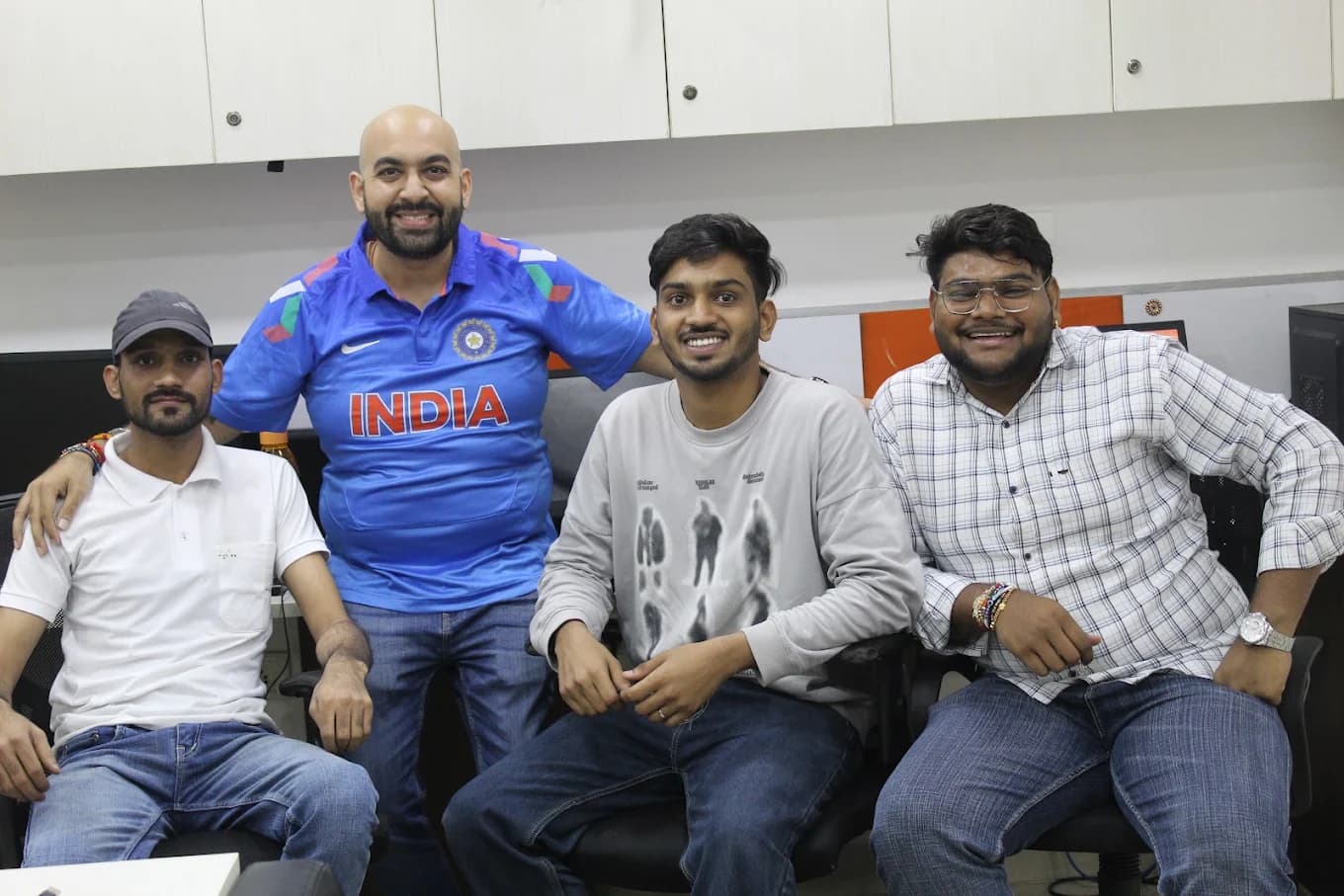 Four colleagues in office chairs at the desks, one in a blue India cricket jersey, arms round each other's shoulders.