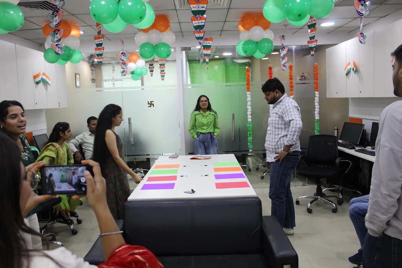 A colleague in a green shirt at the head of the boardroom table laid with coloured paper squares for a game, another filming on a phone in the foreground.