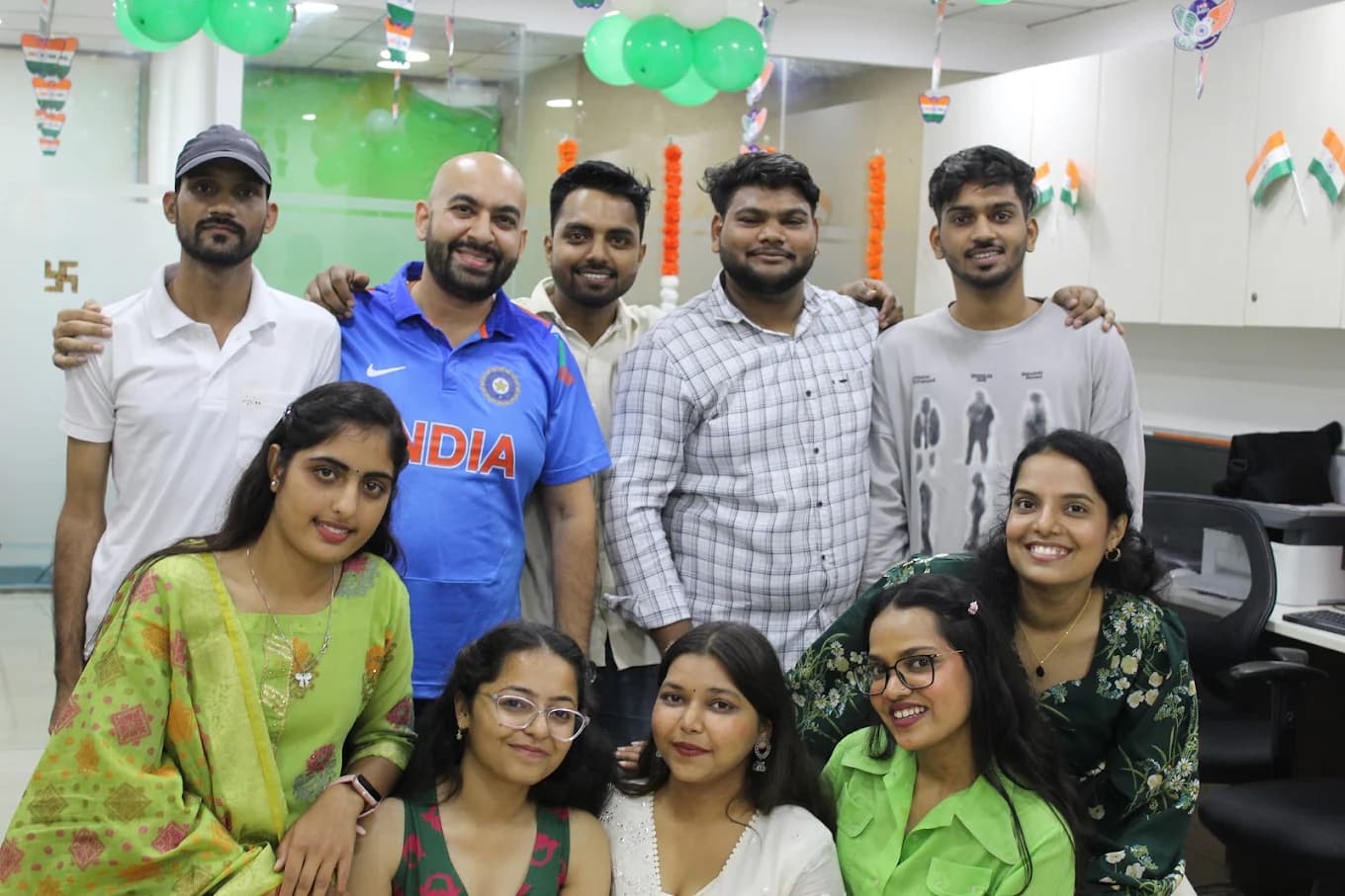 Ten team members, five standing behind five seated, under tricolour balloons and Jai Ho paper hangers in the office for Independence Day 2025.