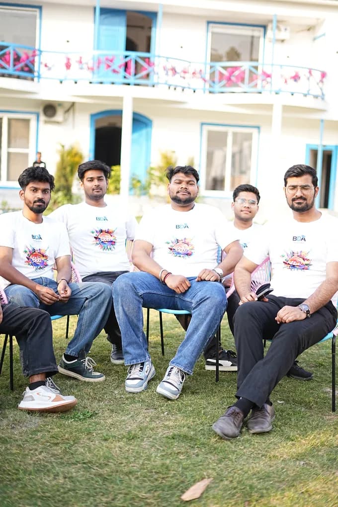 Five colleagues in white Holi t-shirts seated in cane chairs on the lawn in front of the blue-trimmed villa.