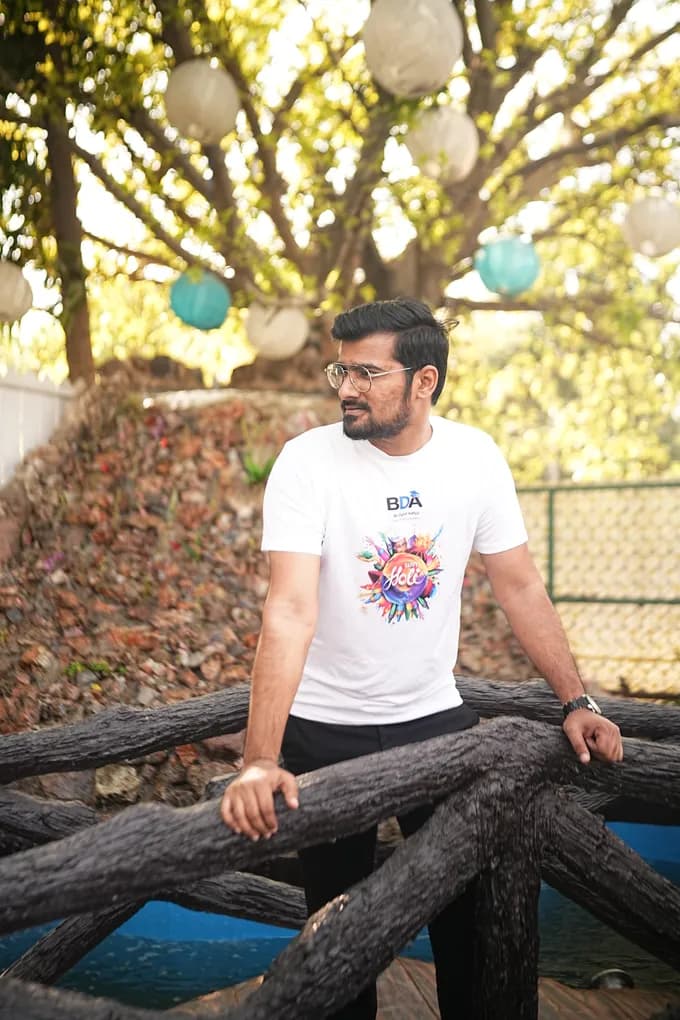A colleague in a white Holi t-shirt leaning on the log railing by the rock wall, paper lanterns hung in the tree above.