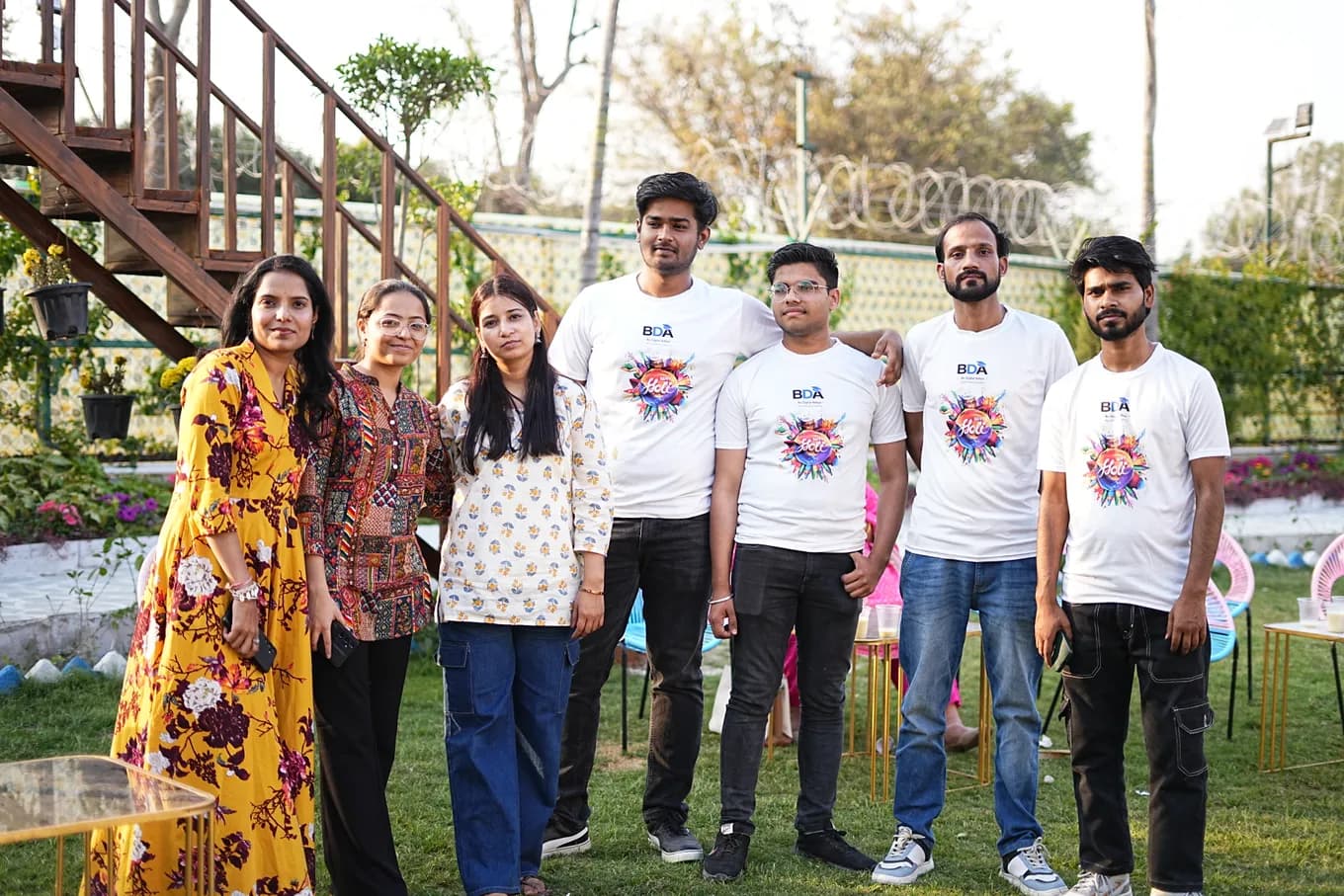 Seven colleagues with arms round each other's shoulders on the lawn in front of the farmhouse stairs, four in white Holi t-shirts.