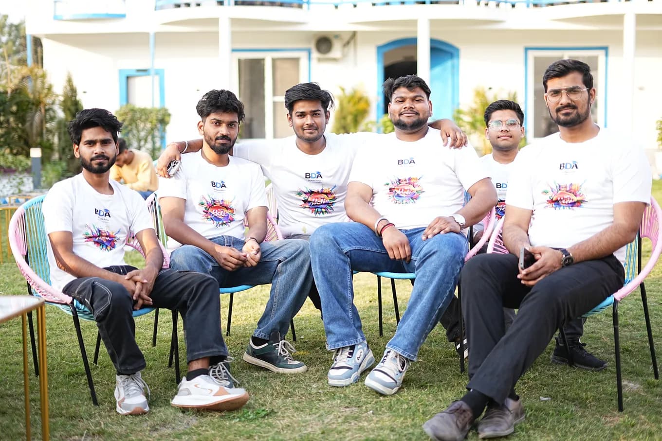 Six colleagues in white Holi t-shirts sitting in a row of pastel cane chairs on the lawn, the blue-trimmed villa behind them.