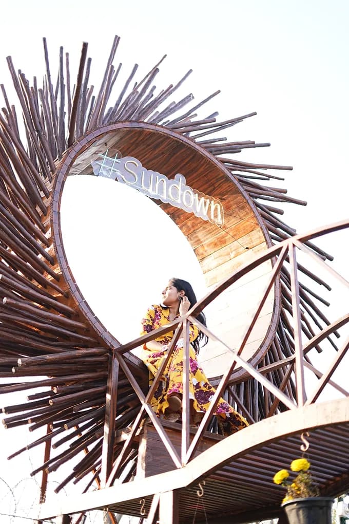 A colleague in a yellow floral dress seated inside the round Sundown sign, a sunburst of wooden sticks above the farmhouse staircase.