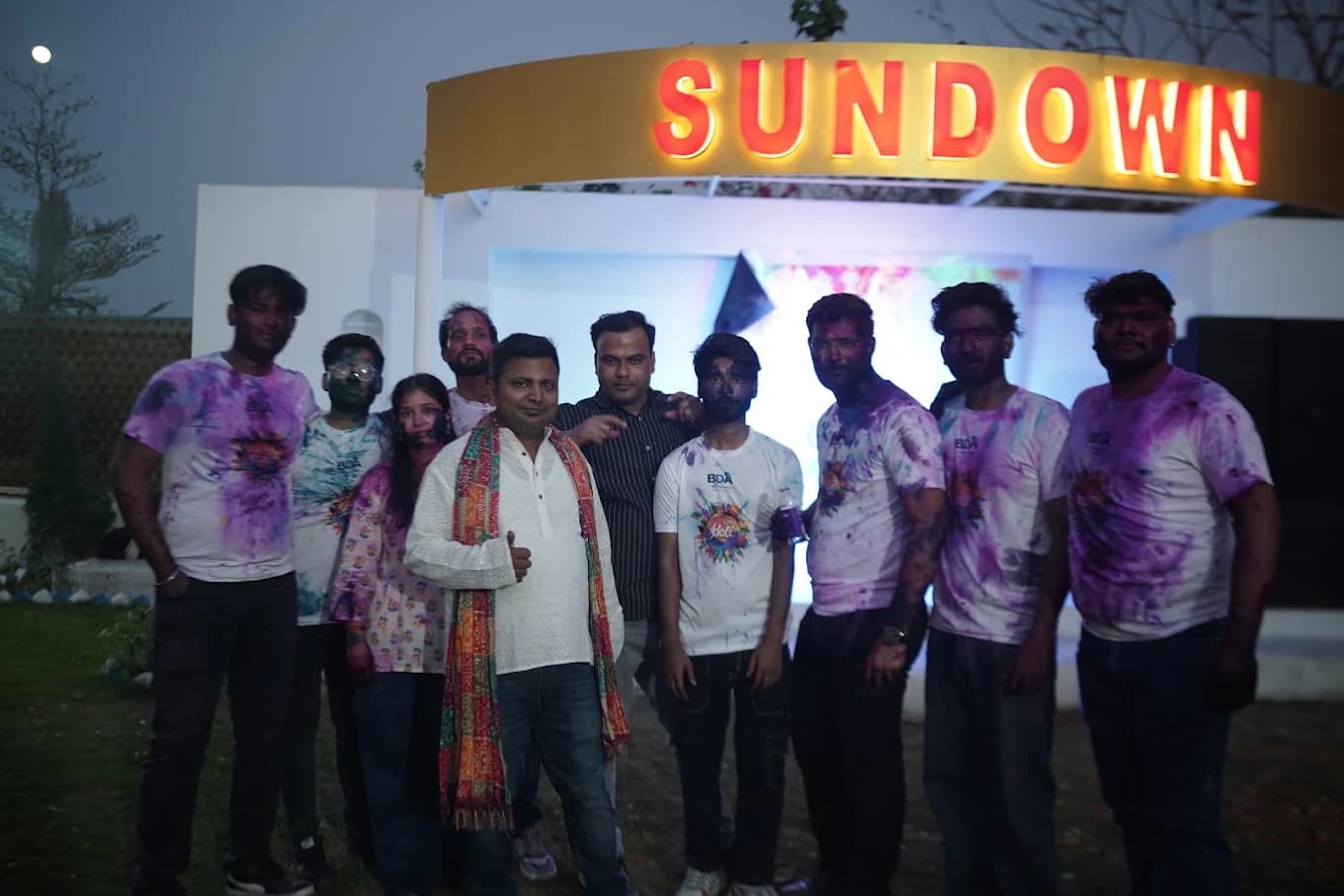Ten team members in colour-splashed white Holi t-shirts, faces smeared with pink and purple gulal, under a lit Sundown sign at dusk.