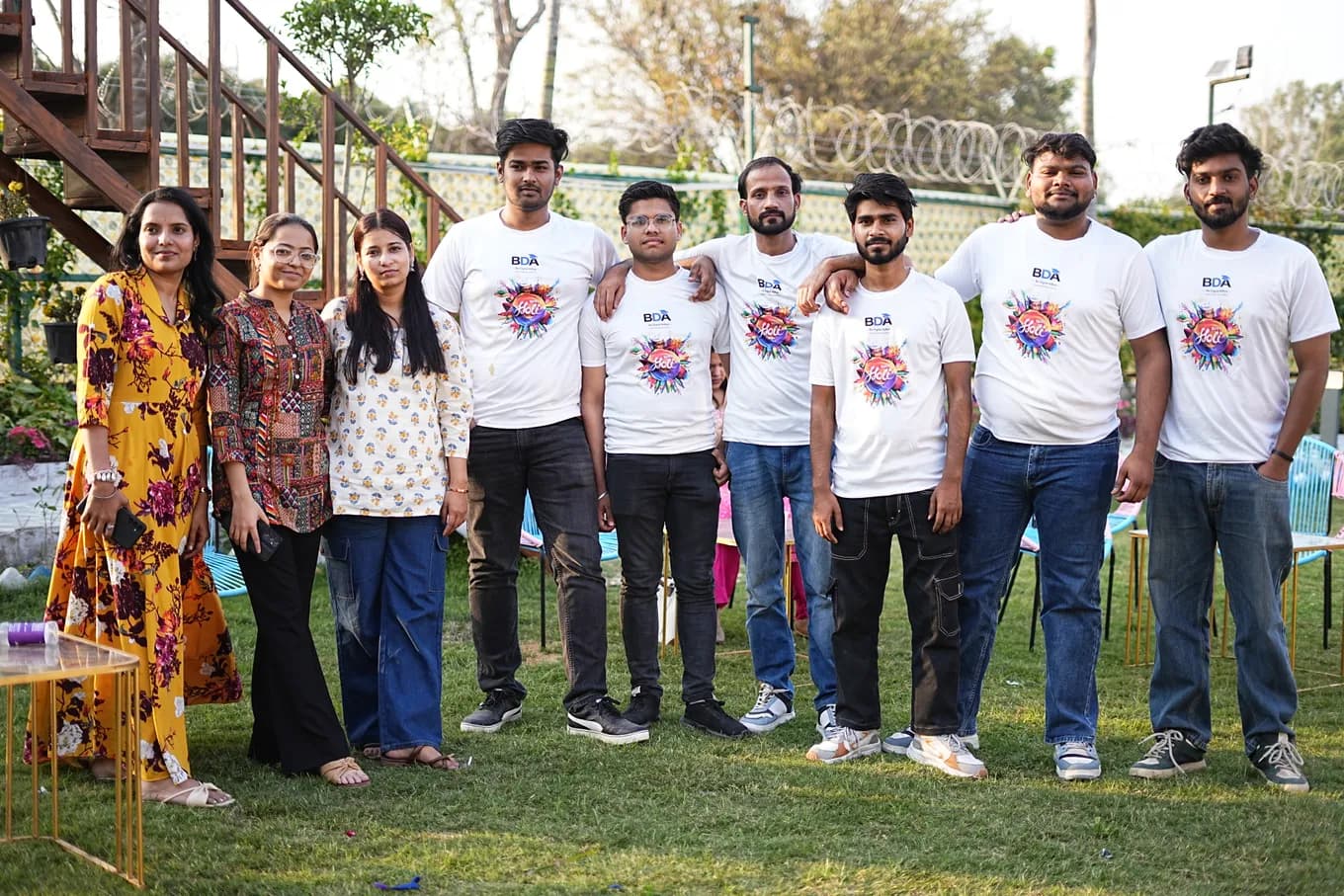 Ten team members in white Holi t-shirts and printed kurtas lined up on the farmhouse lawn under the wooden staircase, before the colour.