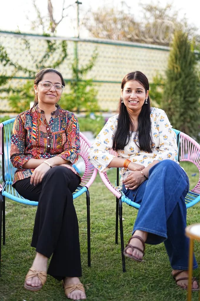 Two colleagues in printed kurtas sitting in pink and blue cane chairs on the farmhouse lawn.