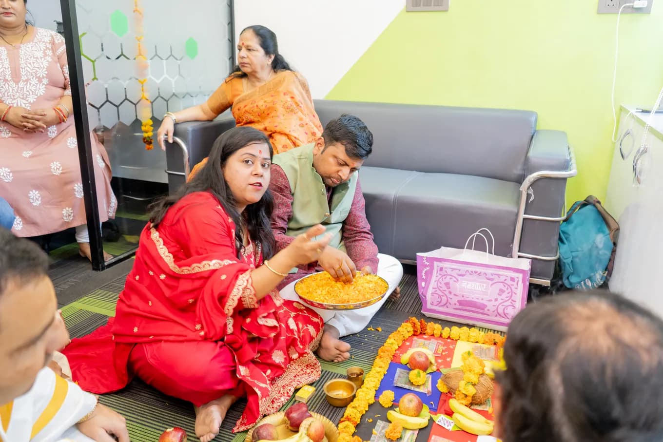 A woman in red and a man in a maroon kurta and green jacket seated at the pooja offerings on the carpet, an elder watching from the sofa behind them.