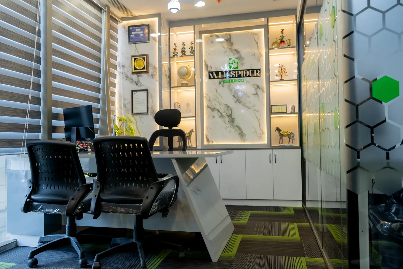 A cabin in the new office with the Web Spider Solutions logo on a marble panel, awards and trophies on backlit shelves, two black chairs and green-striped carpet.