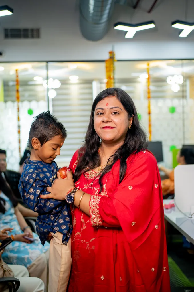 A woman in a red suit holding a small child with an apple in the open workspace, marigold garlands hanging behind them.