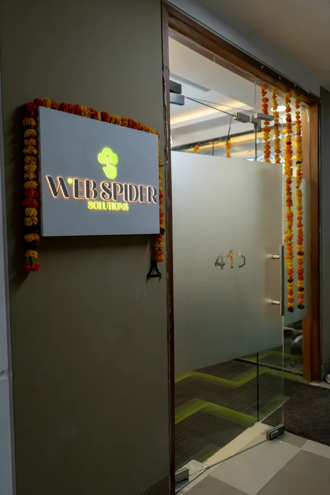 The lit Web Spider Solutions logo sign beside the frosted glass entrance of the new Noida office, a marigold garland hung over it.