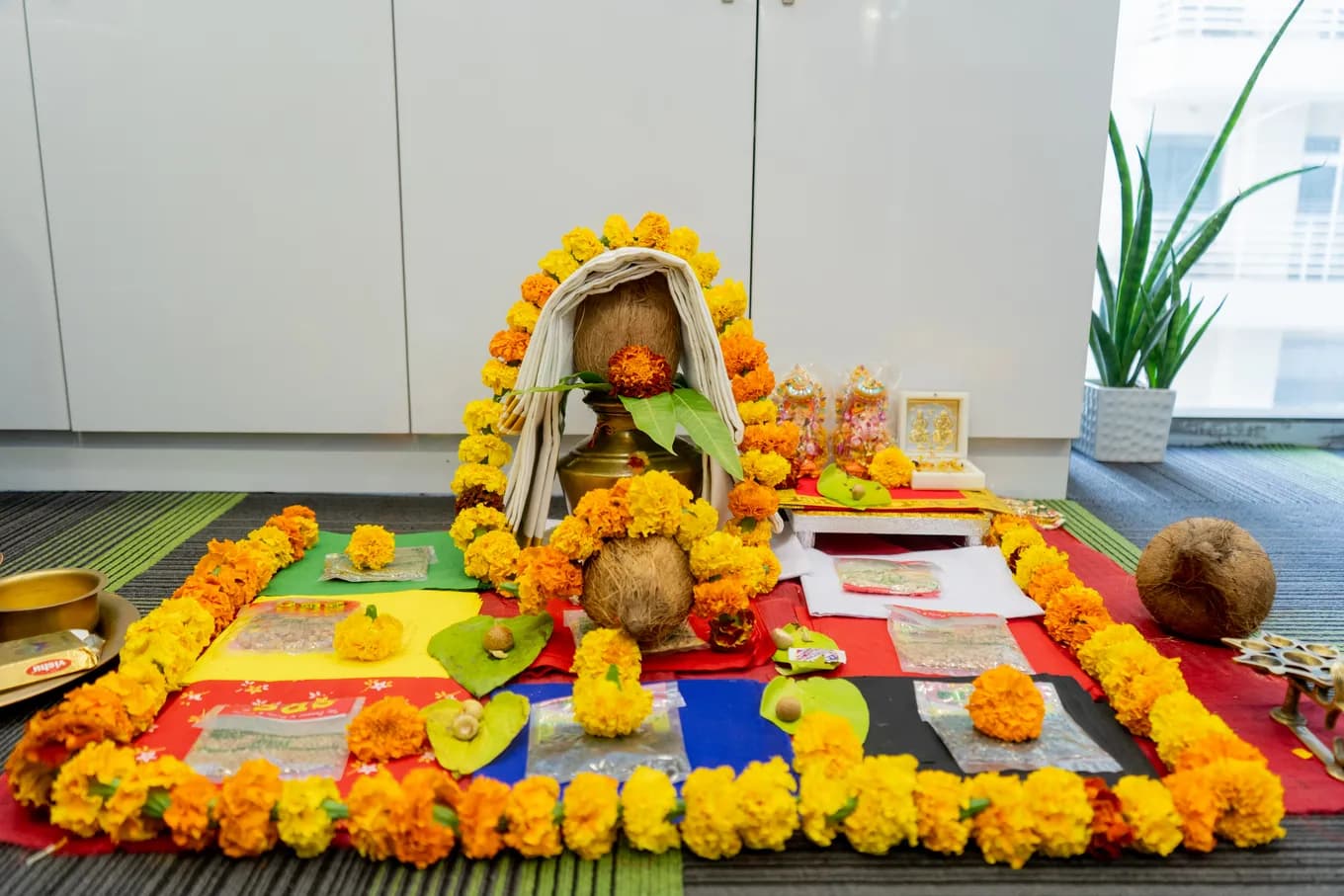 The housewarming pooja laid out on the office carpet: a marigold border, a brass kalash topped with a coconut and mango leaves, a framed idol, coloured cloths and sweets.