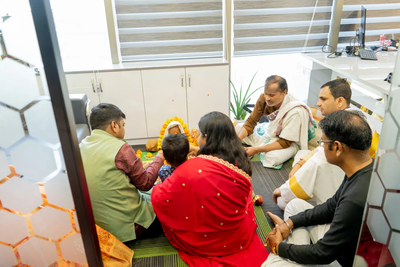 Family and colleagues seated on the carpet with the priest in white for the housewarming pooja, seen through the hexagon-patterned glass partition.