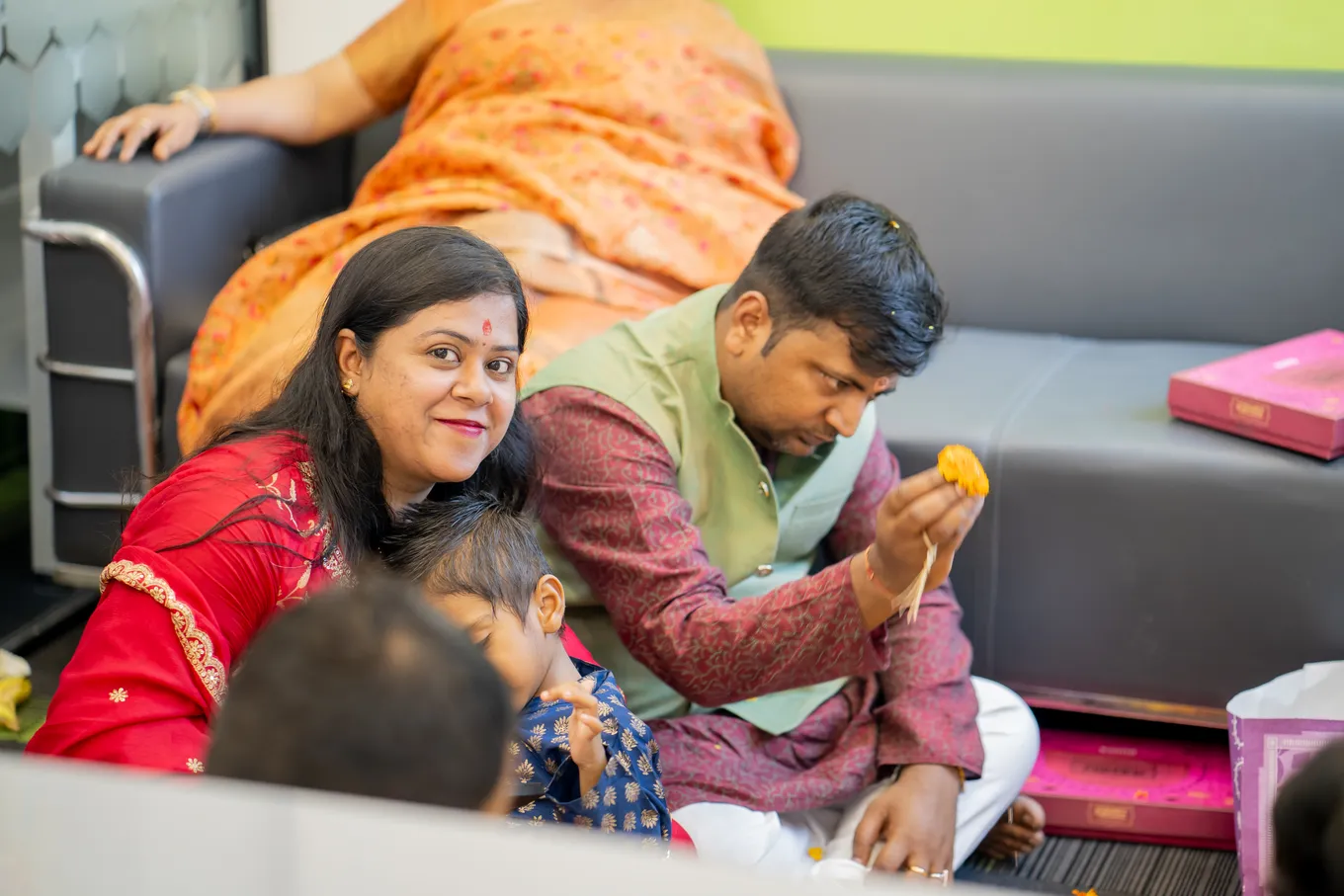 A couple on the carpet after the pooja, the man holding out a laddoo to a small child while the woman smiles at the camera.