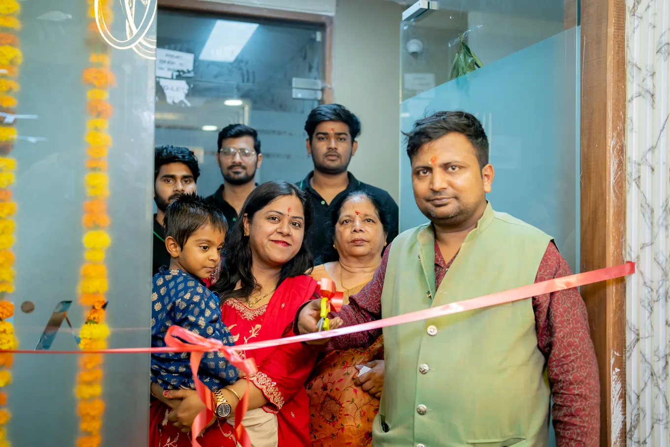 A woman in red cutting the red ribbon across the garlanded glass door of the new office, family and colleagues holding it on either side.