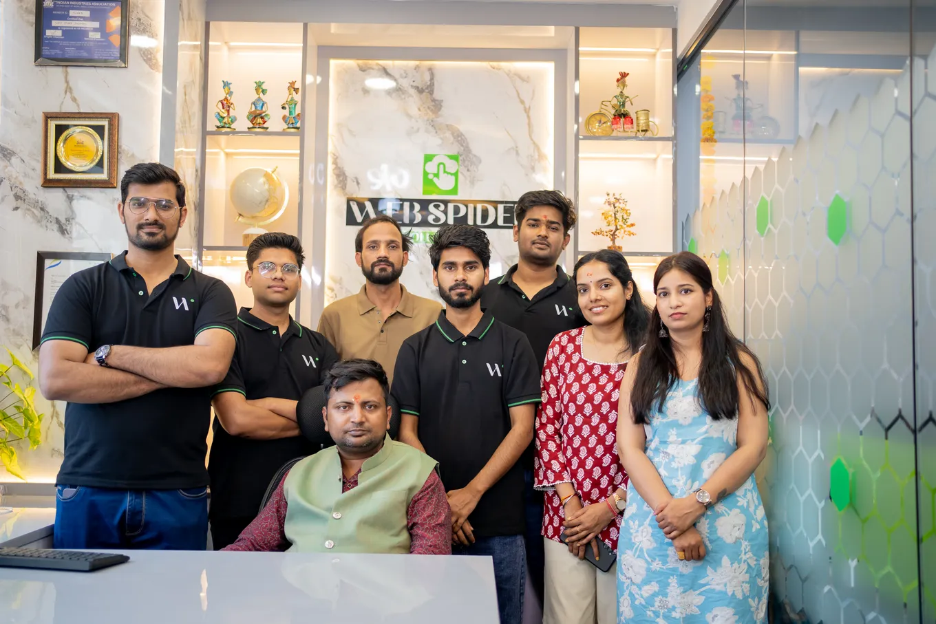 The whole team gathered around the founder's chair in front of the Web Spider Solutions logo wall in the new cabin, most in black polos.