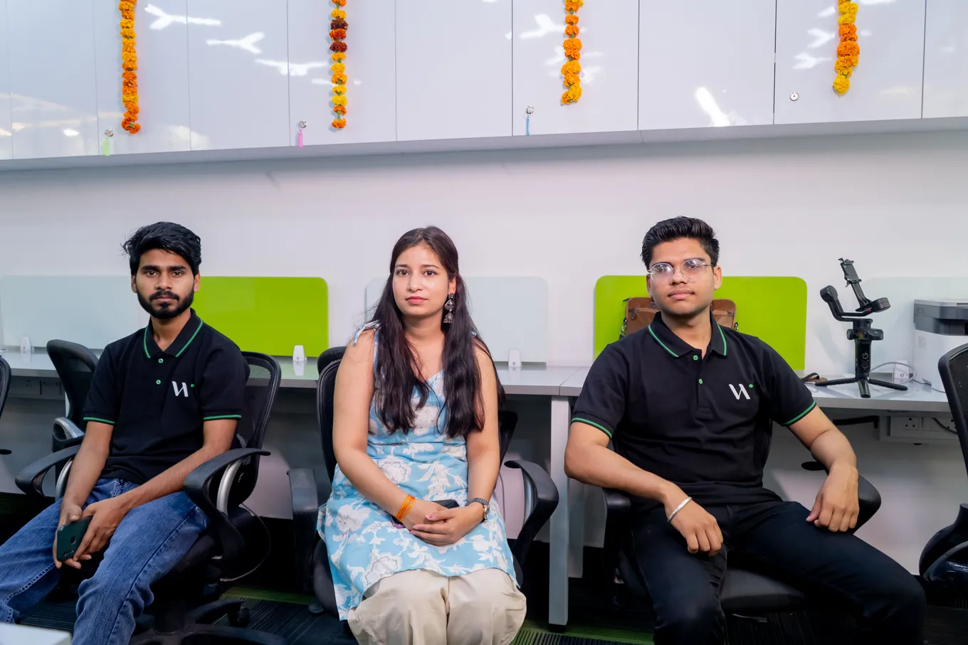 Three colleagues seated at the new desks, two in black Web Spider polos and one in a blue floral dress, marigold strings hanging above.