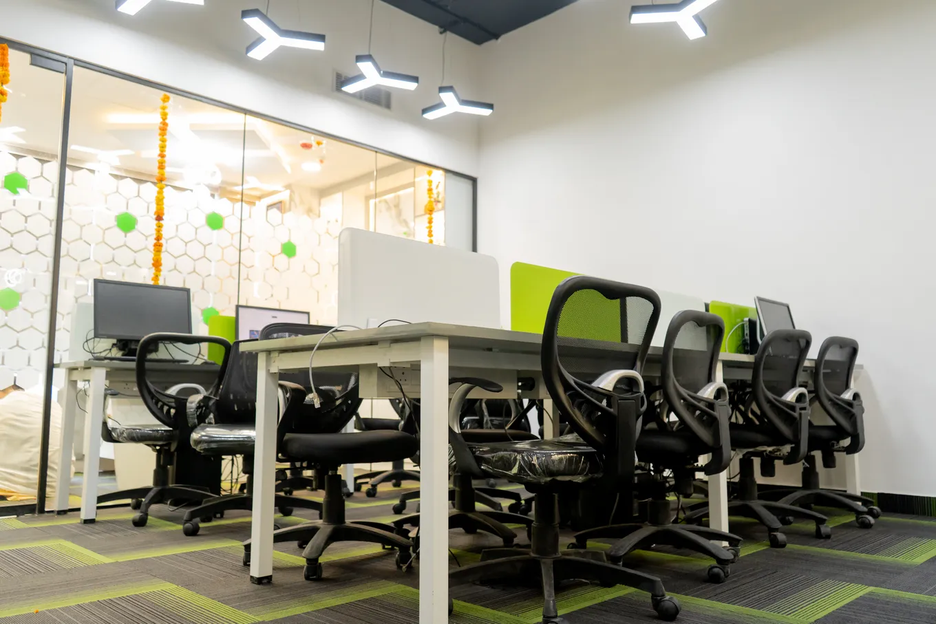 The open workspace of the new office: white desks with black chairs and green panels under Y-shaped ceiling lights, marigold garlands on the glass partitions.
