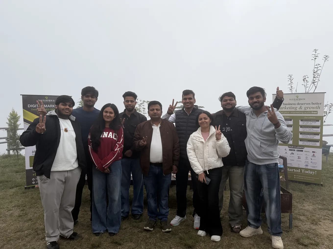 Nine team members flashing peace signs between two Web Spider Solutions roll-up banners on a misty hilltop at Chakrata.