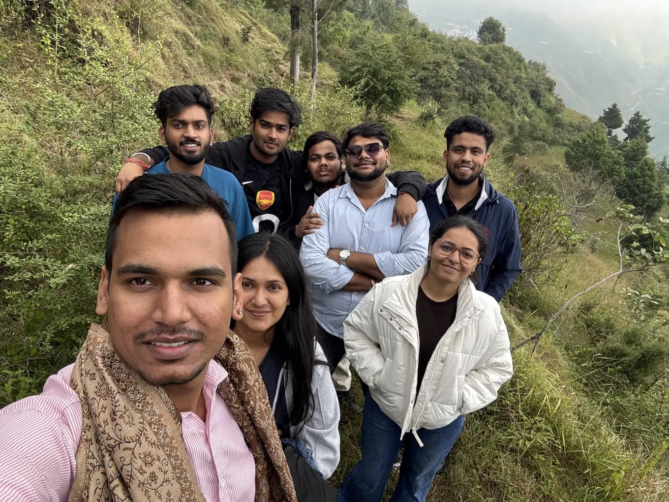 Group selfie of eight team members on a grassy hillside path at Chakrata, forested slopes falling away behind.