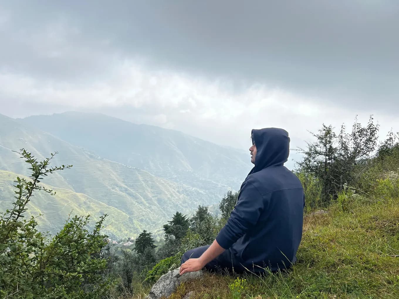 A colleague in a navy hoodie sitting on the grass, looking out over cloud-covered terraced hills.