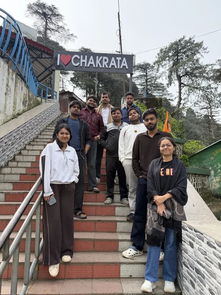 Eight colleagues on a red and white staircase under an I love Chakrata sign, deodar trees behind them.