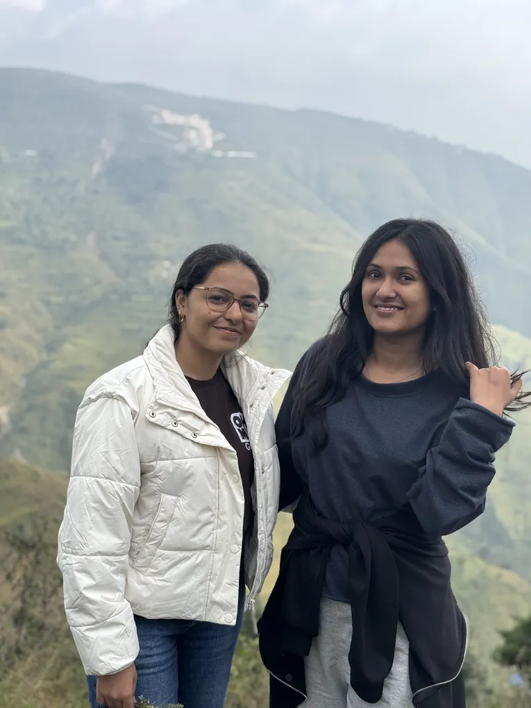 Two colleagues, one in a white puffer jacket and one in a navy sweatshirt, smiling in front of a green terraced mountainside.