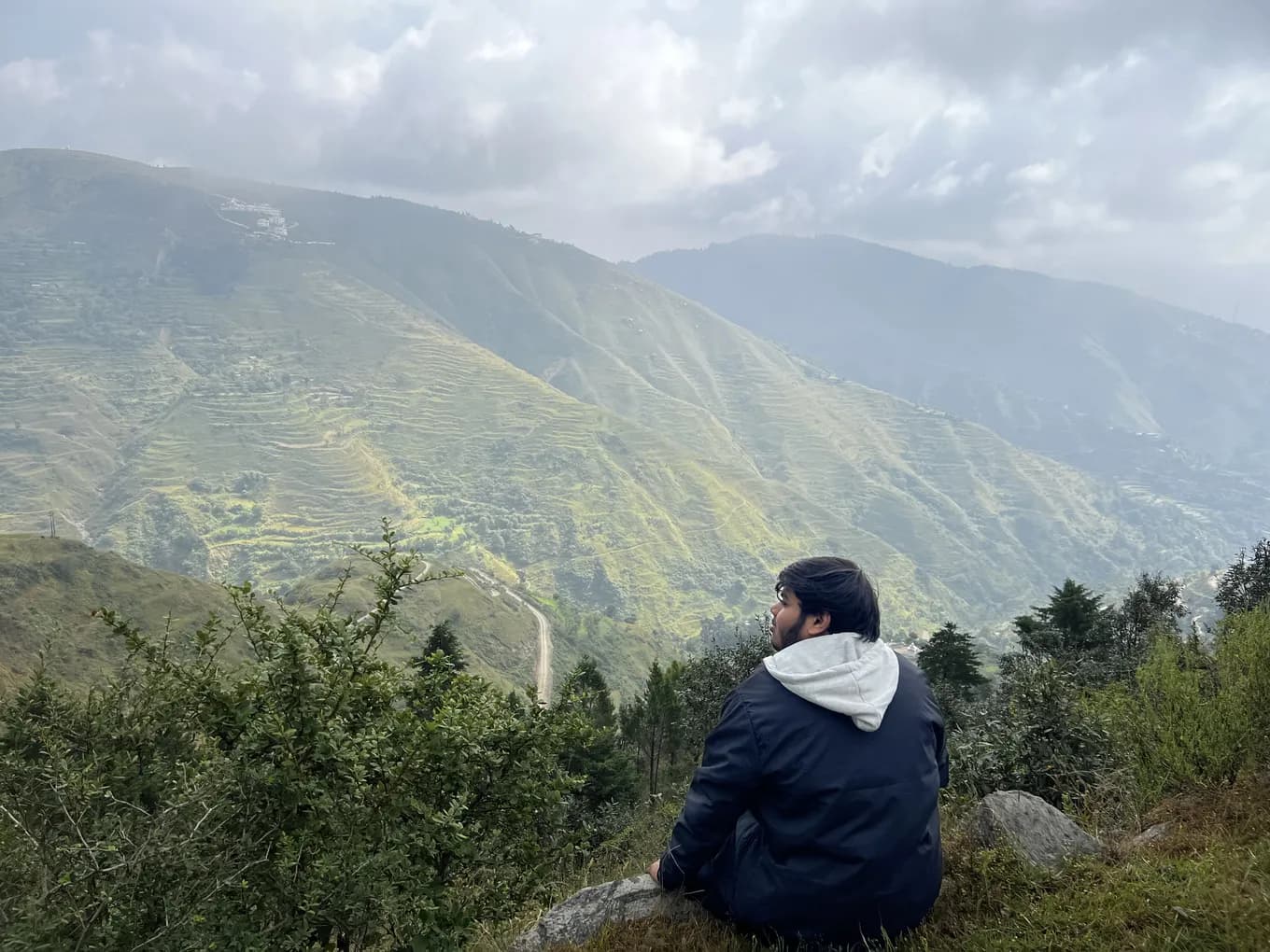 A colleague in a hooded jacket sitting on a rock, looking across to terraced slopes and a hill road at Chakrata.