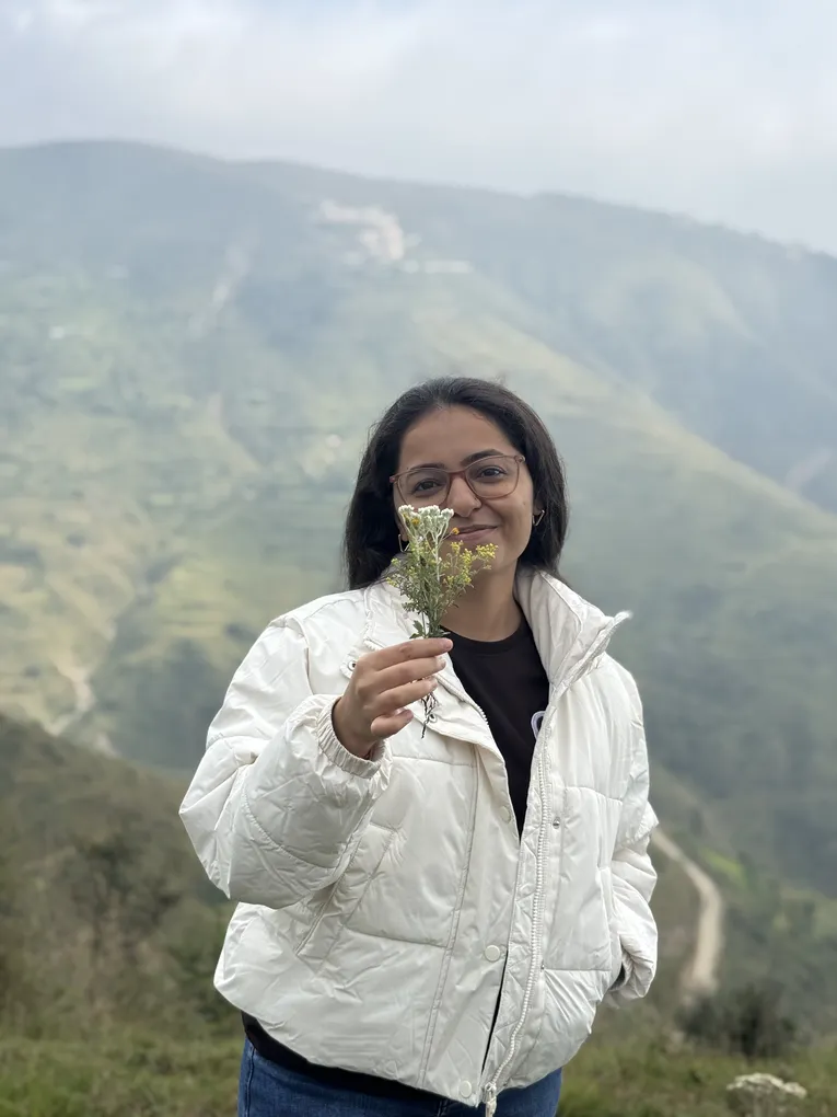 A colleague in a white puffer jacket holding a sprig of white wildflowers, terraced hills behind her.